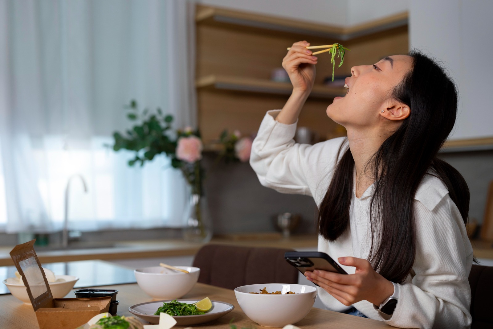 seorang perempuan sedang makan sayur sambil memegang HP dan memakai baju putih