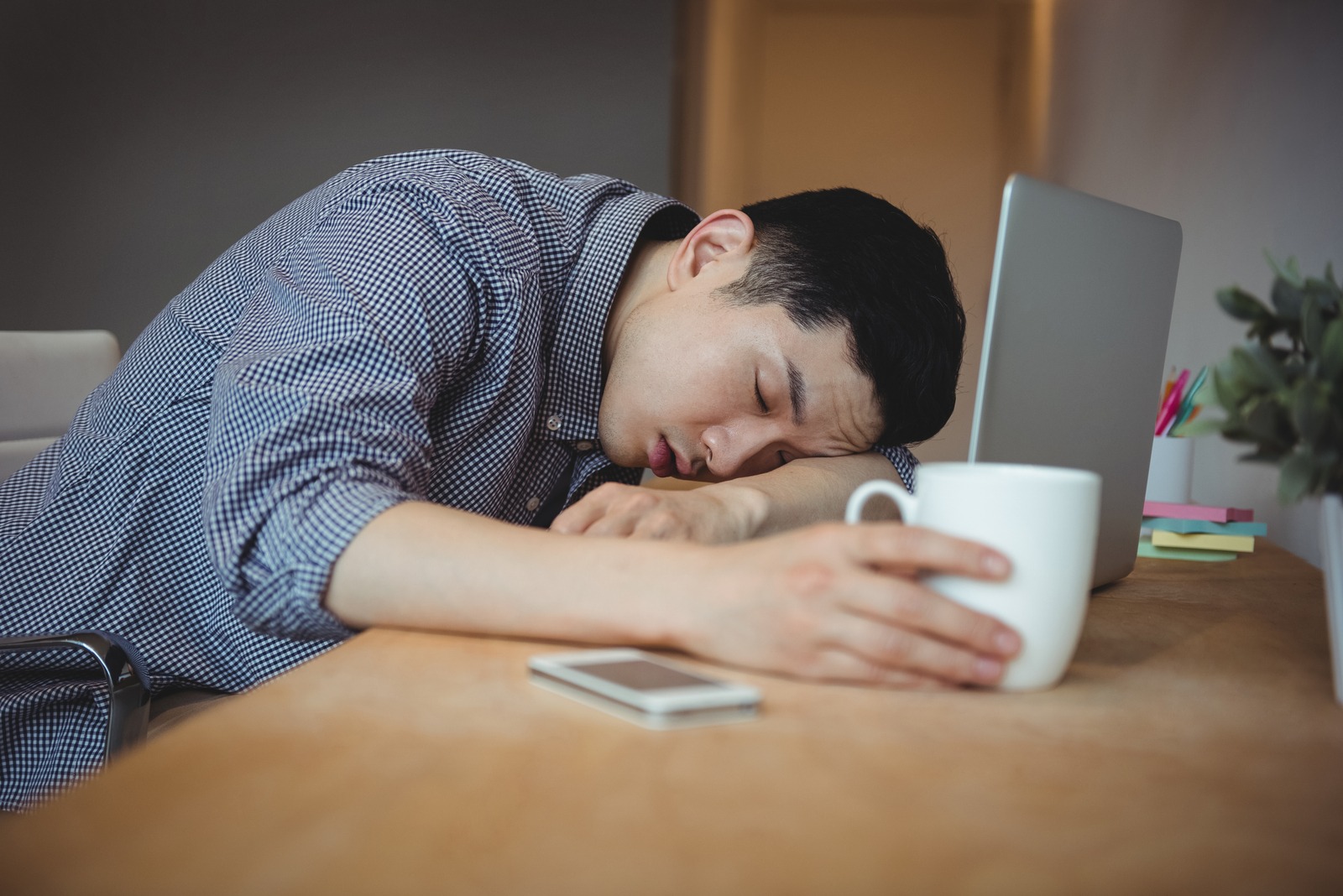 Seorang pria sedang tertidur di atas laptop sambil memegang gelas putih