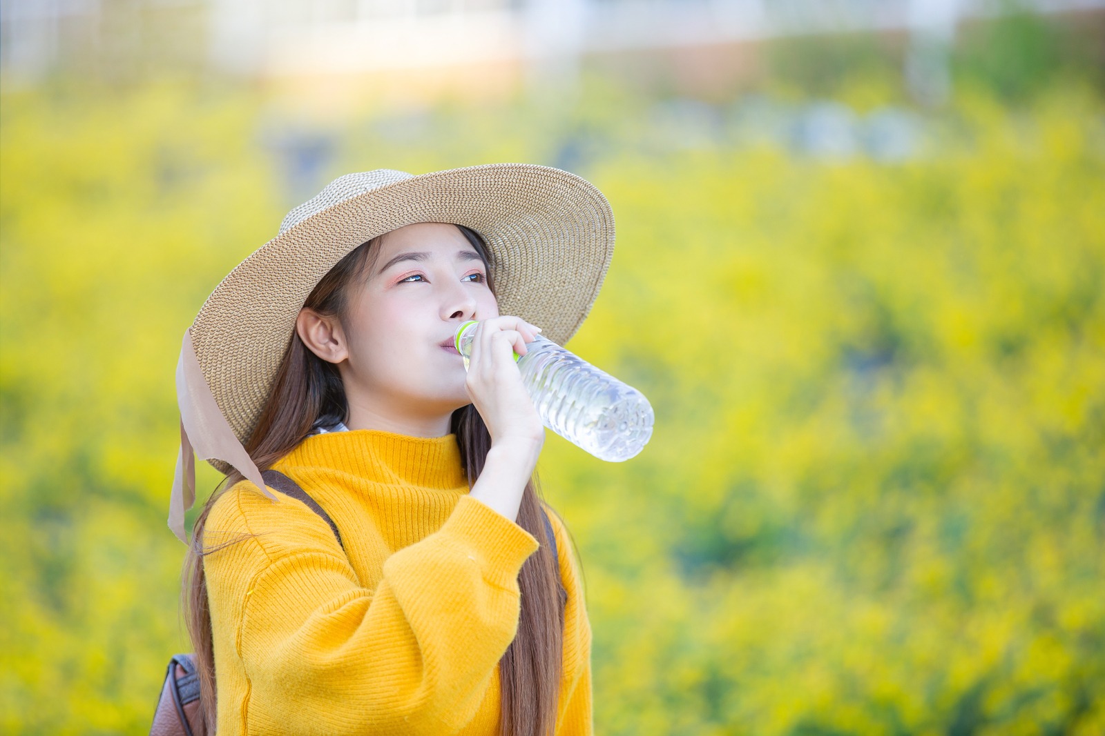 wanita sedang meminum sebotol air mineral memakai baju kuning dan topi besar