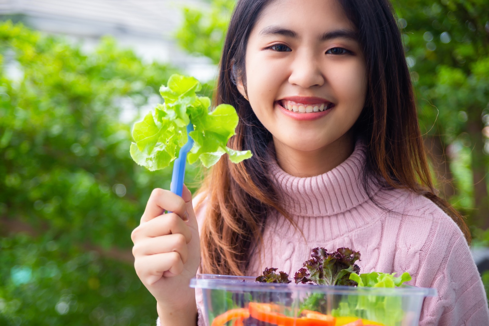 Perempuan pakai baju pink sedang makan sayuran sambil tersenyum